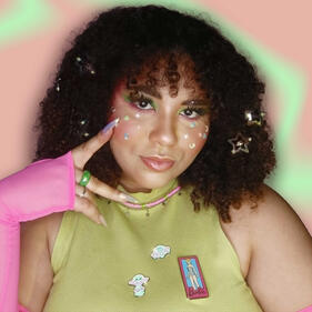 Mulher de pele parda, cabelo cacheado castanho longo com penteado, maquiagem leve com delineado gráfico branco e estrelas rosas e verdes, usando blusa de manga longa branca, com as mãos na cabeça em frente a fundo verde e rodeada pela palavra "lunática"